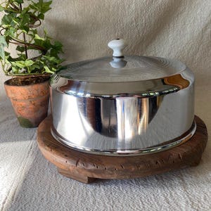 May include: A round, polished chrome cake cover with a white ceramic knob handle, resting on a circular, dark brown, textured wooden base. The metal dome features subtle horizontal grooved lines near the top, reflecting light across its surface.