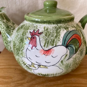 May include: A ceramic teapot with a green lid and handle. The body of the teapot is white with a green speckled pattern and a painted rooster design. The rooster has red, white, and green accents.