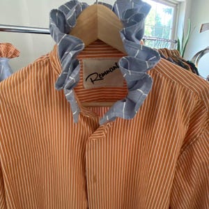 Peut inclure: Une chemise boutonn&eacute;e &agrave; rayures verticales orange et blanches dot&eacute;e d'un col &agrave; volants bleu et blanc &agrave; motif quadrill&eacute;. L'&eacute;tiquette int&eacute;rieure au niveau du cou affiche le texte Remnant en police cursive noire sur un &eacute;cusson beige.