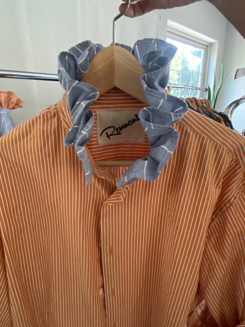 Peut inclure: Une chemise boutonn&eacute;e &agrave; rayures verticales orange et blanches dot&eacute;e d'un col &agrave; volants bleu et blanc &agrave; motif quadrill&eacute;. L'&eacute;tiquette int&eacute;rieure au niveau du cou affiche le texte Remnant en police cursive noire sur un &eacute;cusson beige.