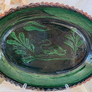 May include: An oval, handmade ceramic platter with a dark green and black glaze. The platter features a decorative design of green leaves and a bird. The rim has a textured, scalloped edge with a reddish-brown color.