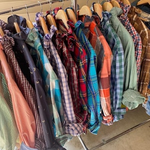 May include: A collection of long-sleeved button-down shirts hanging on wooden hangers on a metal rack. The shirts feature various patterns including plaid, gingham, and solid colors such as orange, blue, red, green, and brown, with some shirts displaying small embroidered animal motifs.