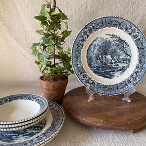 May include: A set of ceramic dinnerware featuring a blue and white transferware pattern. The collection includes a large plate on a stand and a stack of smaller bowls and plates, all decorated with a detailed winter landscape and floral border.