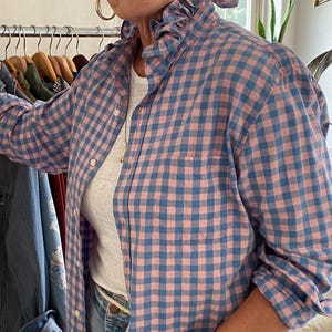 Peut inclure: Une chemise &agrave; manches longues boutonn&eacute;e avec un motif &agrave; carreaux vichy rose et bleu, un col &agrave; volants et une poche poitrine unique. La chemise est port&eacute;e sur un sous-v&ecirc;tement blanc uni et associ&eacute;e &agrave; un jean en denim bleu clair.