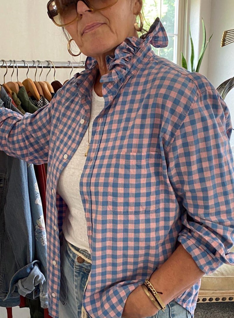 Peut inclure: Une chemise &agrave; manches longues boutonn&eacute;e avec un motif &agrave; carreaux vichy rose et bleu, un col &agrave; volants et une poche poitrine unique. La chemise est port&eacute;e sur un sous-v&ecirc;tement blanc uni et associ&eacute;e &agrave; un jean en denim bleu clair.