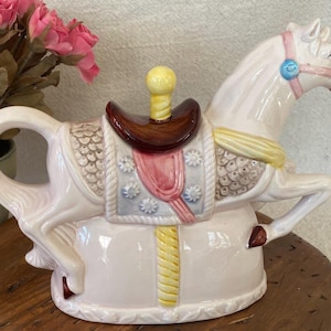 May include: A ceramic carousel horse teapot in white with a brown saddle, pink and blue accents, and a yellow knob on the lid. The teapot has a handle and a spout, and is sitting on a wooden surface.