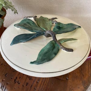 May include: A cream-colored ceramic lid with a raised leaf design in shades of green and gray. The lid is resting on a wooden surface. The lid's rim has a thin gray border. The overall theme is kitchenware.