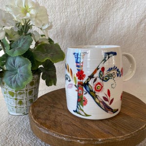 Puede incluir: Taza de cer&aacute;mica blanca con un gran dise&ntilde;o floral "K" colorido. La letra est&aacute; adornada con patrones florales rojos, azules y verdes. La taza tiene un asa curva y se apoya en un soporte de madera. Una planta en maceta con flores blancas y hojas verdes est&aacute; en el fondo.