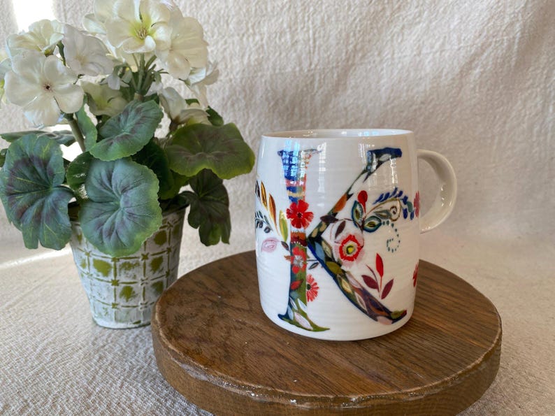 Puede incluir: Taza de cer&aacute;mica blanca con un gran dise&ntilde;o floral "K" colorido. La letra est&aacute; adornada con patrones florales rojos, azules y verdes. La taza tiene un asa curva y se apoya en un soporte de madera. Una planta en maceta con flores blancas y hojas verdes est&aacute; en el fondo.
