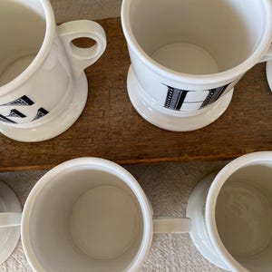 May include: Several white ceramic mugs with black letter designs are displayed. Each mug has a handle and a small, round base. The mugs are arranged on a wooden surface, presenting a simple, classic design.