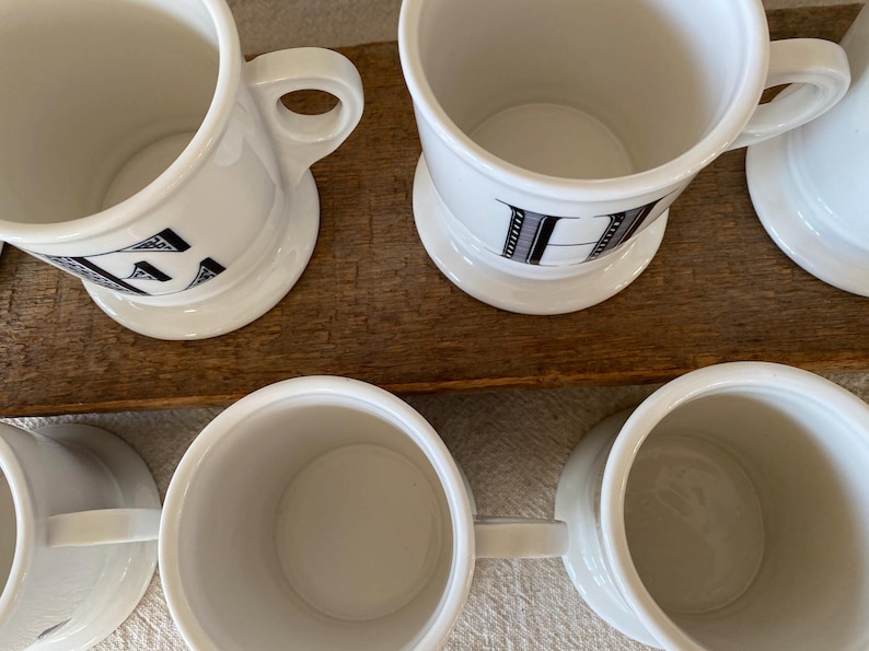 May include: Several white ceramic mugs with black letter designs are displayed. Each mug has a handle and a small, round base. The mugs are arranged on a wooden surface, presenting a simple, classic design.