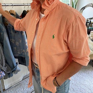 Peut inclure: Une chemise boutonn&eacute;e &agrave; carreaux vichy orange et blanc avec un col &agrave; volants et un petit logo vert brod&eacute; sur la poitrine gauche. La chemise pr&eacute;sente des manches longues, une patte de boutonnage et est port&eacute;e sur un t-shirt blanc avec un jean bleu clair.