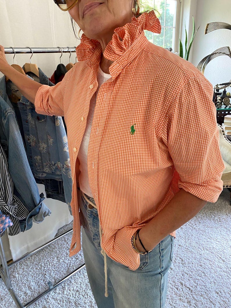 Peut inclure: Une chemise boutonn&eacute;e &agrave; carreaux vichy orange et blanc avec un col &agrave; volants et un petit logo vert brod&eacute; sur la poitrine gauche. La chemise pr&eacute;sente des manches longues, une patte de boutonnage et est port&eacute;e sur un t-shirt blanc avec un jean bleu clair.