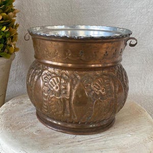 May include: A copper-colored metal planter with a silver rim and handles. The planter features embossed designs of figures and patterns. A small potted plant is visible in the background.