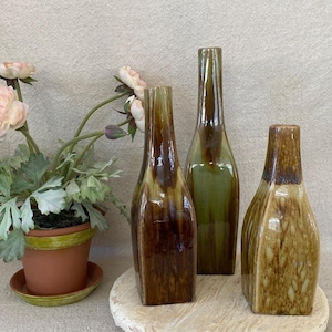 May include: Three tall, slender ceramic vases with a glossy, mottled glaze in shades of brown, tan, and olive green, arranged next to a terracotta pot containing a plant with light pink flowers and green, serrated leaves.