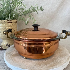 May include: A round copper casserole dish with a matching lid, featuring a black knob handle and two side handles with black accents. The exterior has horizontal ribbed detailing and the lid displays a faint etched botanical design.