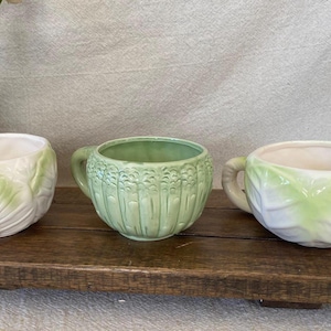 May include: Three ceramic planters in a row, each with a unique design. The left and right planters are white with green leaf patterns. The center planter is light green with a textured vertical design.
