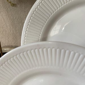 May include: Two white ceramic plates with a ridged, fan-like design around the rims. The plates are stacked, with the top plate partially obscuring the bottom one. The plates are clean and simple, suitable for serving food.