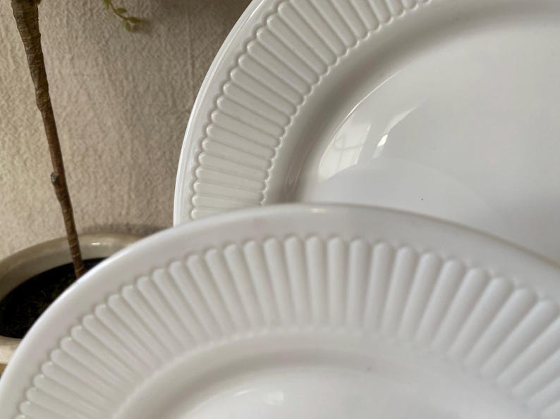 May include: Two white ceramic plates with a ridged, fan-like design around the rims. The plates are stacked, with the top plate partially obscuring the bottom one. The plates are clean and simple, suitable for serving food.