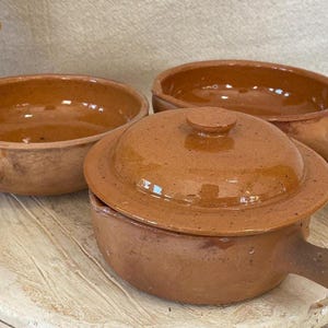 May include: Three rustic, terracotta-colored ceramic bowls with handles. One bowl has a matching lid. The bowls are round with a slightly rough texture and a warm, earthy tone, suggesting handmade craftsmanship.