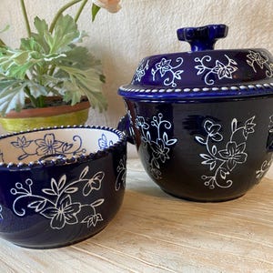 May include: Two-piece ceramic serving set in a deep blue hue, adorned with white floral patterns. The set includes a lidded casserole dish and a smaller bowl, both featuring a beaded rim. The casserole has handles and a decorative knob.