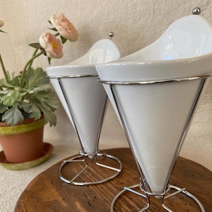 May include: Two white ceramic cone-shaped serving dishes with a silver metal frame. The dishes are on a wooden surface, with a potted plant with pink flowers in the background. The dishes have a small silver ball on top.