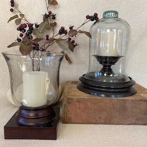 May include: Two glass candle holders with candles, one under a glass cloche. The candle holders are on wooden bases. A sprig of berries and leaves is in one of the candle holders. The cloche is on a wooden block.