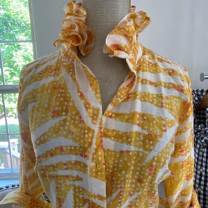 Peut inclure: Une chemise &agrave; manches longues boutonn&eacute;e pr&eacute;sentant un motif abstrait jaune et blanc avec de petits points roses. La chemise poss&egrave;de un col haut &agrave; volants et est pr&eacute;sent&eacute;e sur un mannequin. Le tissu a un aspect textur&eacute; avec des poignets.