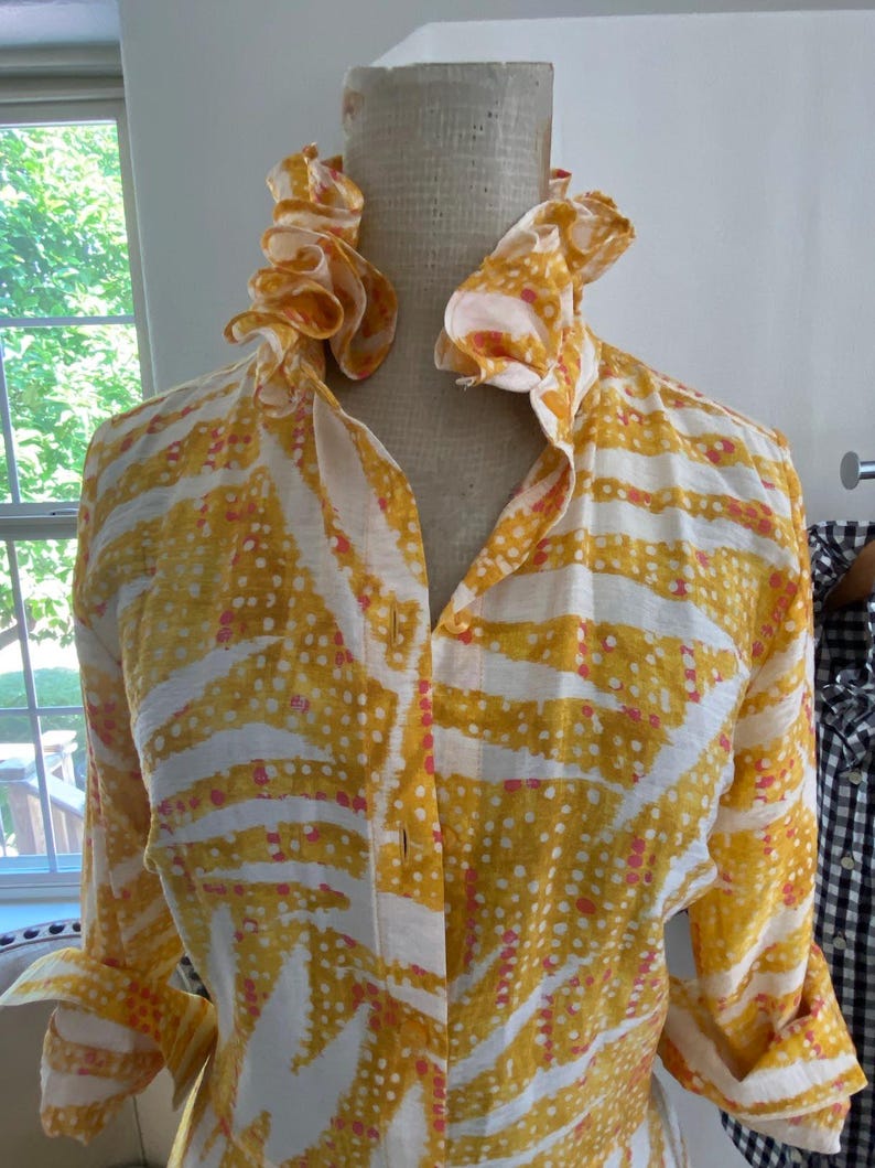 Peut inclure: Une chemise &agrave; manches longues boutonn&eacute;e pr&eacute;sentant un motif abstrait jaune et blanc avec de petits points roses. La chemise poss&egrave;de un col haut &agrave; volants et est pr&eacute;sent&eacute;e sur un mannequin. Le tissu a un aspect textur&eacute; avec des poignets.