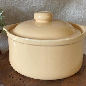 May include: A light beige ceramic casserole dish with a matching lid. The dish has two small handles and a rounded knob on the lid. The casserole dish is sitting on a wooden surface.