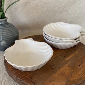 May include: A set of four white ceramic seashell-shaped dishes, stacked on a wooden surface. One dish is separate, showcasing the detailed shell design. A gray vase with greenery is in the background, adding a touch of nature to the scene.