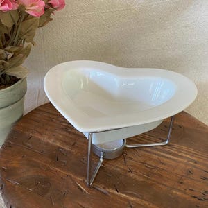 May include: A white ceramic heart-shaped bowl resting on a silver-toned metal wire stand. The stand features a circular holder underneath the bowl, designed to accommodate a small tea light candle for warming the contents of the dish.
