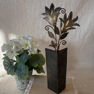 May include: A decorative metal flower arrangement in a dark brown square vase. The flowers have a detailed, open design with small dot accents. A potted plant with white flowers and green leaves is to the left.