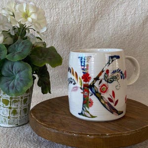 Puede incluir: Taza de cer&aacute;mica blanca con un gran dise&ntilde;o floral "K" colorido. La taza est&aacute; sobre una bandeja redonda de madera, junto a una planta en maceta con flores blancas y hojas verdes. La taza tiene un asa.