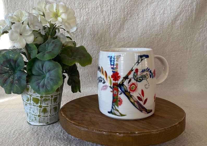 Puede incluir: Taza de cer&aacute;mica blanca con un gran dise&ntilde;o floral "K" colorido. La taza est&aacute; sobre una bandeja redonda de madera, junto a una planta en maceta con flores blancas y hojas verdes. La taza tiene un asa.