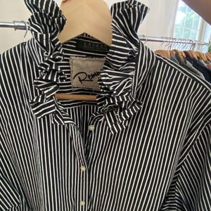 Peut inclure: Une chemise boutonn&eacute;e &agrave; rayures verticales noires et blanches dot&eacute;e d'un col &agrave; volants pro&eacute;minent. Le v&ecirc;tement pr&eacute;sente une &eacute;tiquette avec le texte Lauren Ralph Lauren et un &eacute;cusson avec le texte Remm, tous deux visibles au niveau du col.
