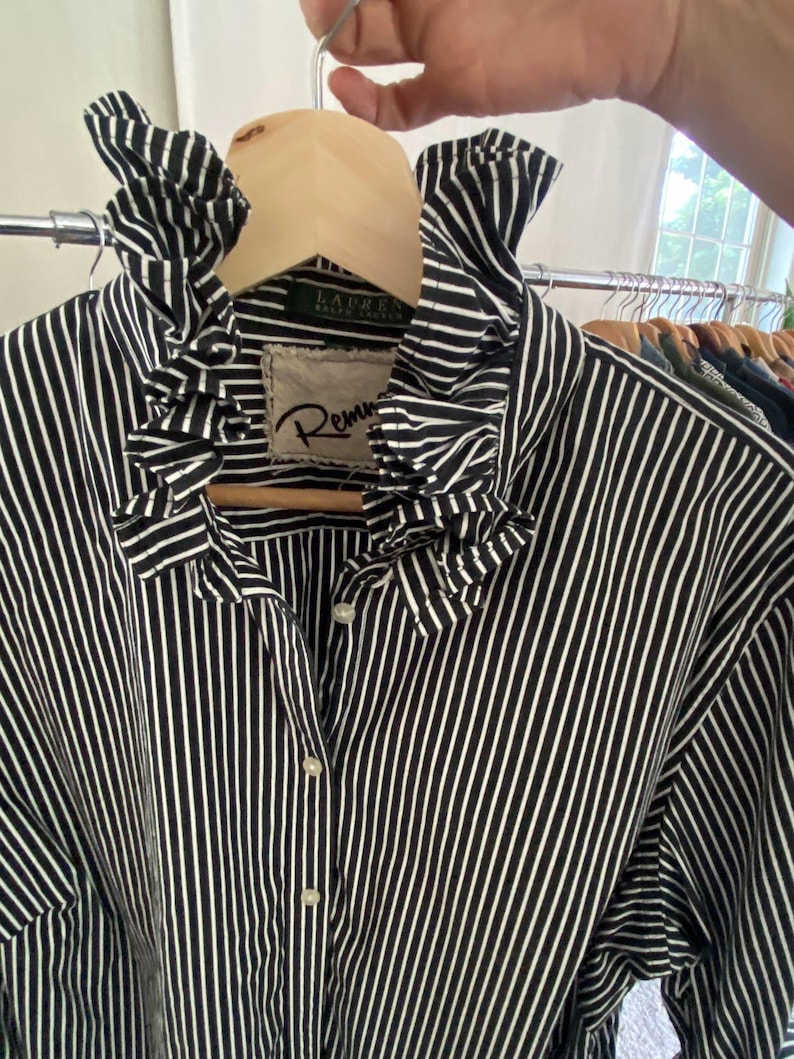 Peut inclure: Une chemise boutonn&eacute;e &agrave; rayures verticales noires et blanches dot&eacute;e d'un col &agrave; volants pro&eacute;minent. Le v&ecirc;tement pr&eacute;sente une &eacute;tiquette avec le texte Lauren Ralph Lauren et un &eacute;cusson avec le texte Remm, tous deux visibles au niveau du col.