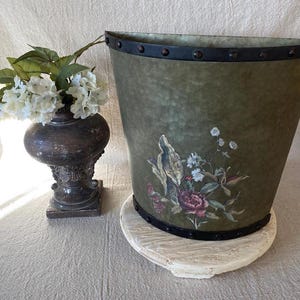 May include: An olive green decorative wastebasket with a floral design, set on a white, round base. The wastebasket has a dark metal trim and rivets. A dark brown vase with white flowers is to the left.