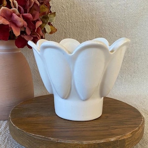 May include: A white ceramic flower-shaped vase with a wide opening. The vase is sitting on a round wooden stand. A peach-colored vase and a bouquet of red flowers are in the background.