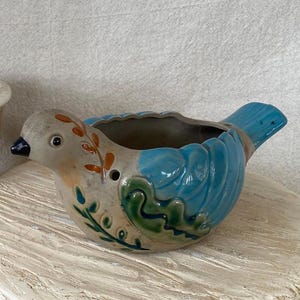 May include: A ceramic bird-shaped planter with a light gray body, blue wings and tail, and a black beak. The bird has orange and green decorative accents. The planter sits on a white, textured surface.