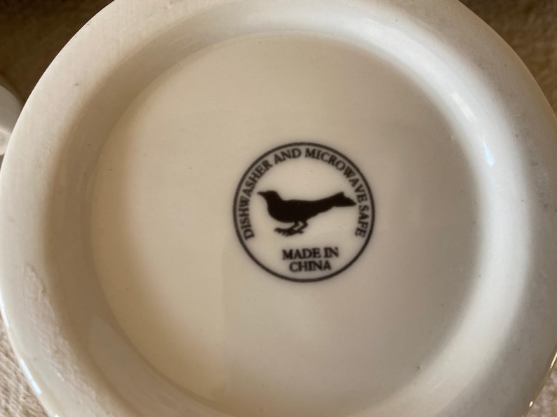May include: A white ceramic mug base with a black circular emblem. The emblem features a bird silhouette and the text "Dishwasher and Microwave Safe" and "Made in China".