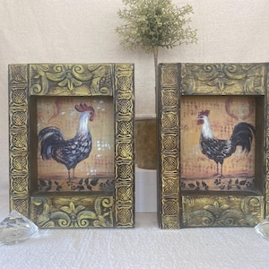 May include: Two framed rooster prints. Each print features a rooster with a red comb, white neck, and black and white body. The frames are gold and black with ornate detailing. Decorative accents are in the foreground.