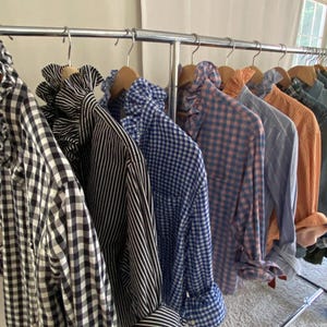 Peut inclure: Une rang&eacute;e de chemises &agrave; manches longues boutonn&eacute;es suspendues sur un portant en m&eacute;tal. Les chemises pr&eacute;sentent divers motifs, notamment du vichy noir et blanc, des rayures verticales noires et blanches, du vichy bleu et blanc et du plaid.