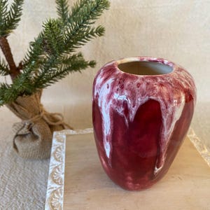 May include: A glossy, red ceramic vase with white dripped glaze. The vase has a rounded shape and a wide opening. A small decorative evergreen tree is in the background.