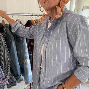 Peut inclure: Une chemise boutonn&eacute;e bleu clair &agrave; rayures verticales blanches, dot&eacute;e d'un col &agrave; volants distinctif dans un tissu ray&eacute; orange et blanc contrastant. La chemise pr&eacute;sente des manches longues, une poche avant et des boutons clairs sur le devant.