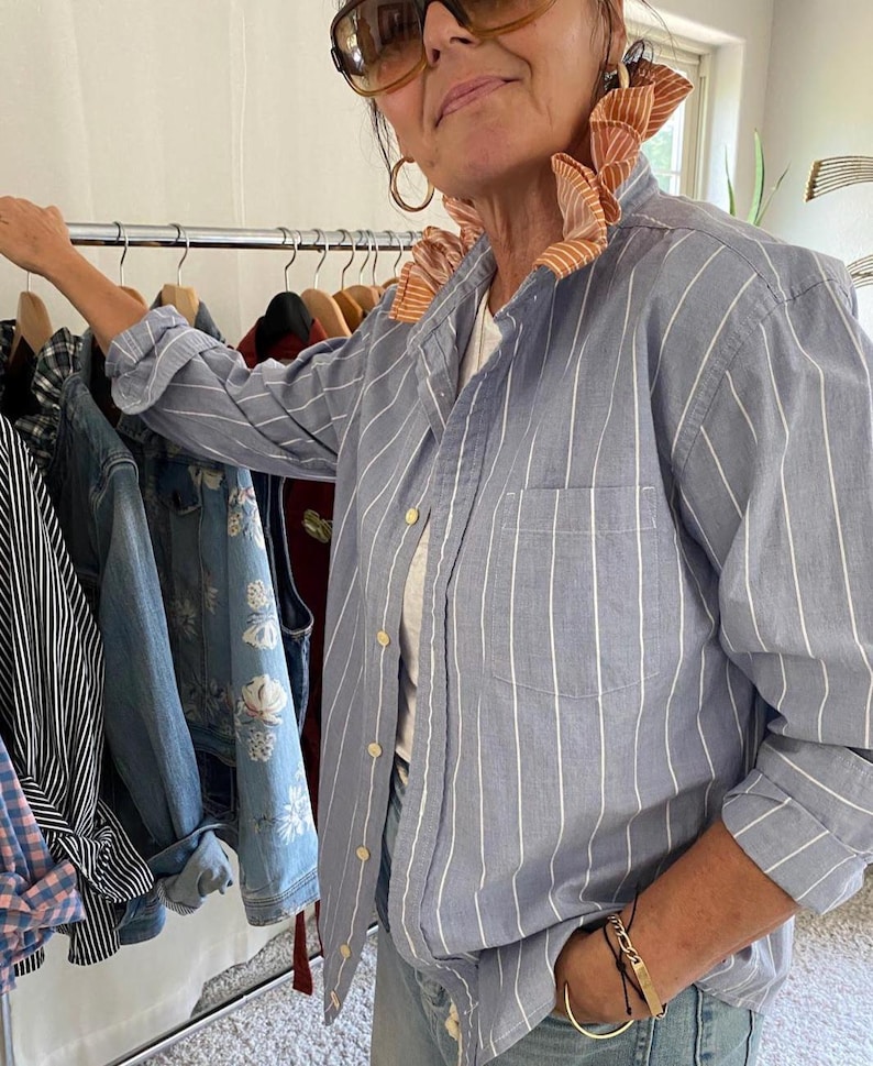 Peut inclure: Une chemise boutonn&eacute;e bleu clair &agrave; rayures verticales blanches, dot&eacute;e d'un col &agrave; volants distinctif dans un tissu ray&eacute; orange et blanc contrastant. La chemise pr&eacute;sente des manches longues, une poche avant et des boutons clairs sur le devant.
