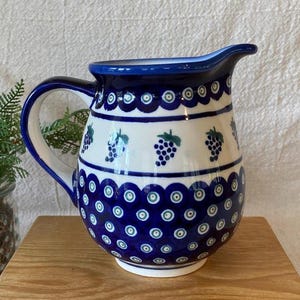 May include: A ceramic pitcher with a white body and a blue handle and spout. The pitcher is decorated with blue and green grape clusters and a repeating blue and white pattern. The pitcher is sitting on a wooden surface.