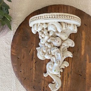 May include: Ornate, off-white wall shelf with a fruit and floral design, mounted on a round, dark brown wooden plaque. The shelf features a decorative edge and detailed carvings, creating a vintage aesthetic. The wood has a rustic, distressed appearance.