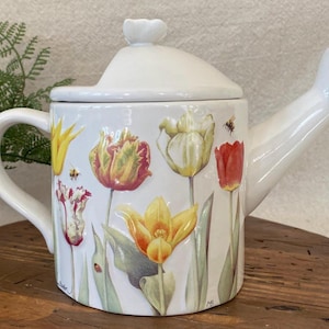 May include: White ceramic watering can with a lid and handle, decorated with colorful tulip illustrations and small bees. The spout is angled, and the handle is curved. The lid has a small flower-shaped knob.