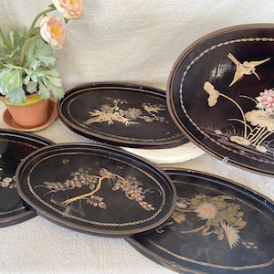 May include: A set of five oval black lacquer trays featuring intricate gold-toned illustrations of birds, flowers, and foliage. Each tray is decorated with unique botanical and avian motifs, including lotus blossoms and flowering branches, with a thin gold border.